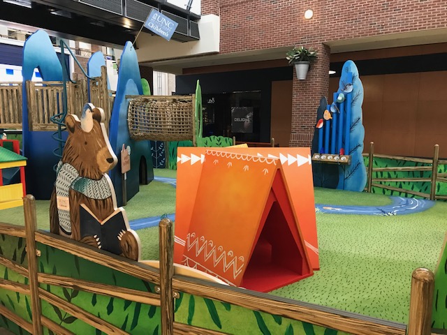 Custom printed indoor playground carpeting with an outdoor camping print that meets ASTM F1292 standards. There is a play ground, an orange tent and a bear reading a book inside this indoor playground facility.